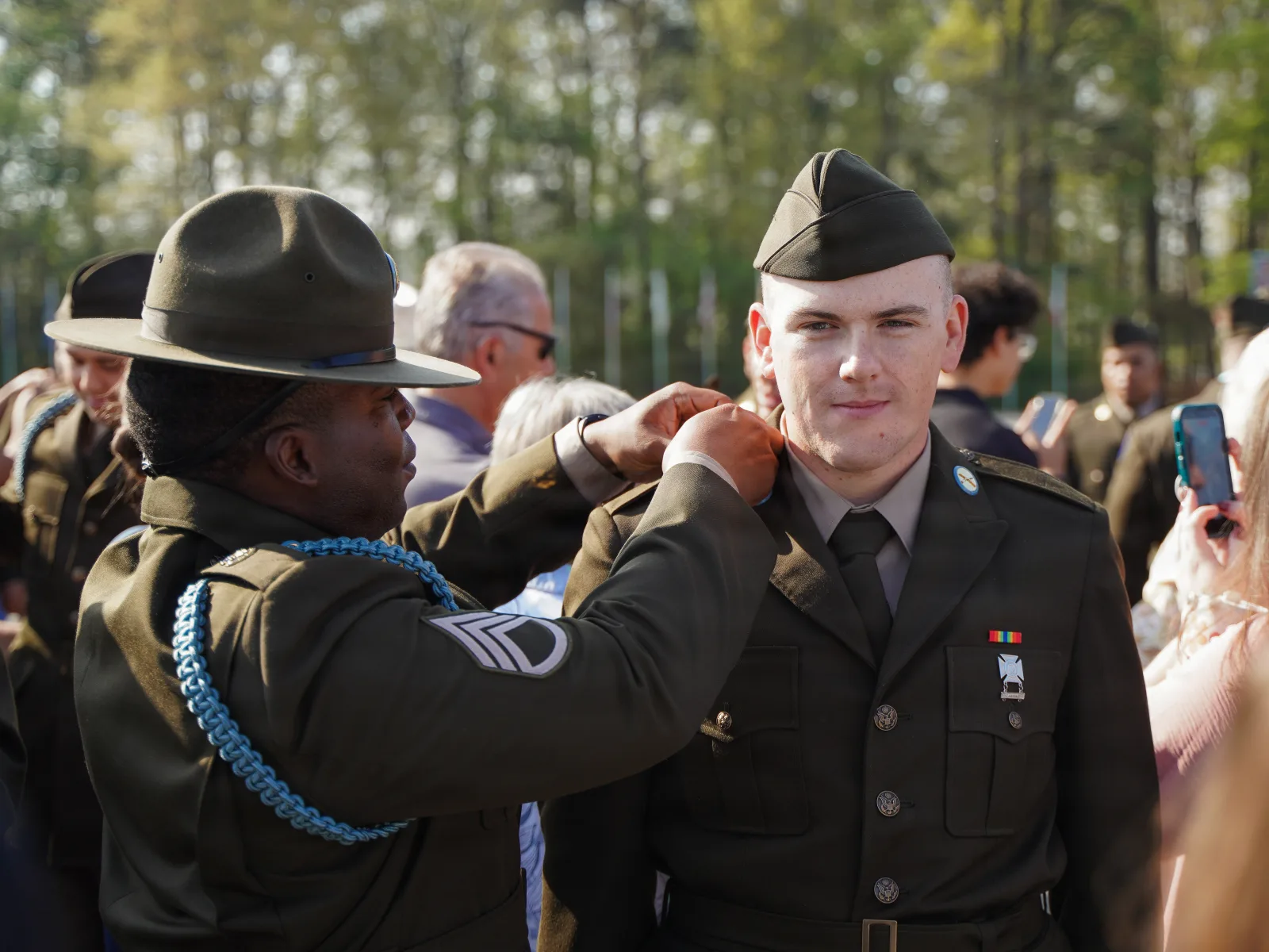 A drill sergeant loops the Infantry blue cord onto a new Soldier's shoulder during a Turning Blue ceremony at Pomeroy Field, Fort Benning, March 2026. U.S. Army photo via DVIDS.